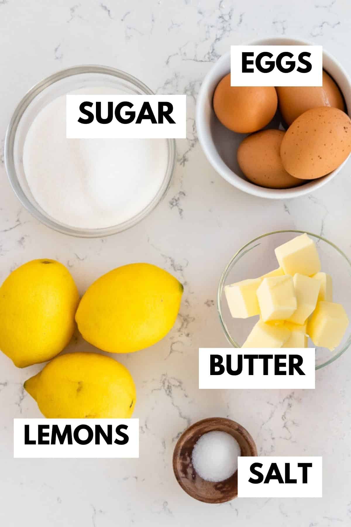 lemons, a bowl of sugar, a bowl of eggs, a bowl of butter and a wood bowl of salt on counter.