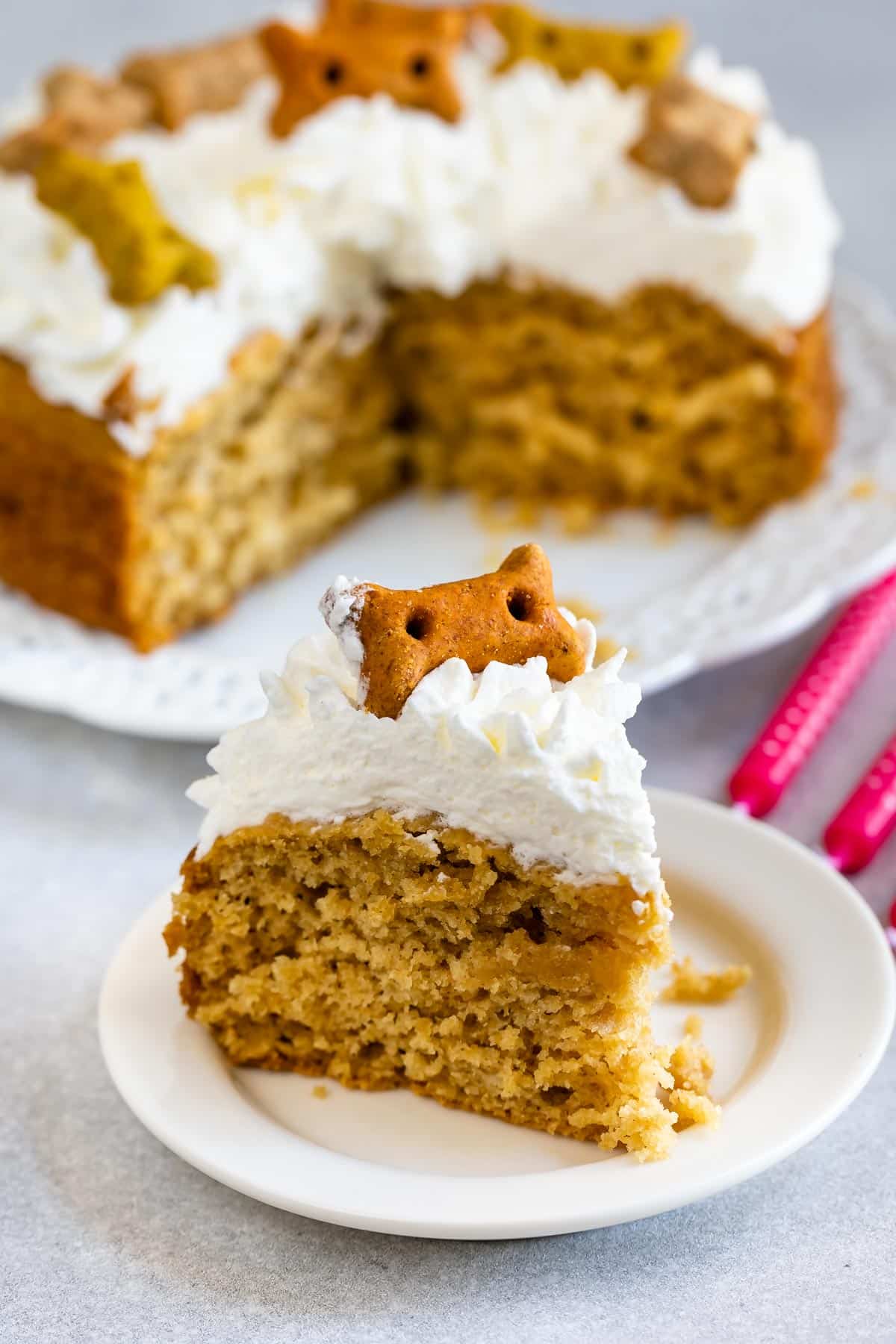 A slice of dog cake with white frosting and a dog-shaped biscuit on top, served on a small white plate in front of the remaining cake and two pink forks.