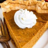 A slice of easy pumpkin pie topped with a swirl of whipped cream sits on a white plate next to a fork engraved with the words YOUR PIE. The pie has a golden, flaky crust.