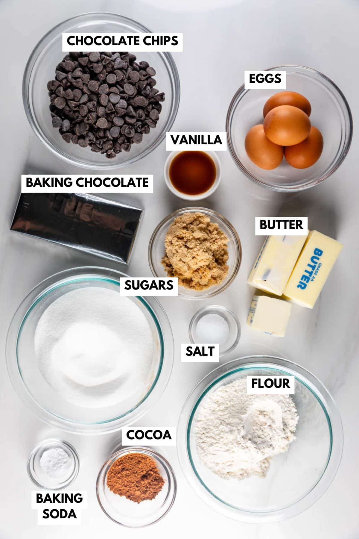Ingredients for baking brookies are arranged on a white surface. They include labeled glass bowls of chocolate chips, eggs, brown and white sugar, vanilla, baking chocolate, butter, salt, flour, cocoa, and baking soda.
