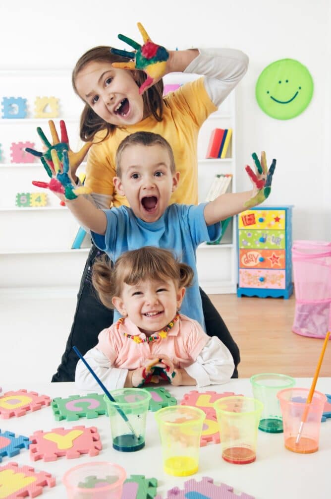 three children in a vertical row holding up hands with paint on them