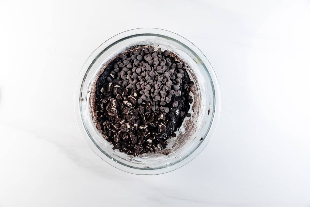 A glass bowl filled with rich chocolate batter, crushed Chocolate Oreo Cookies, and chocolate chips sits on a white surface.