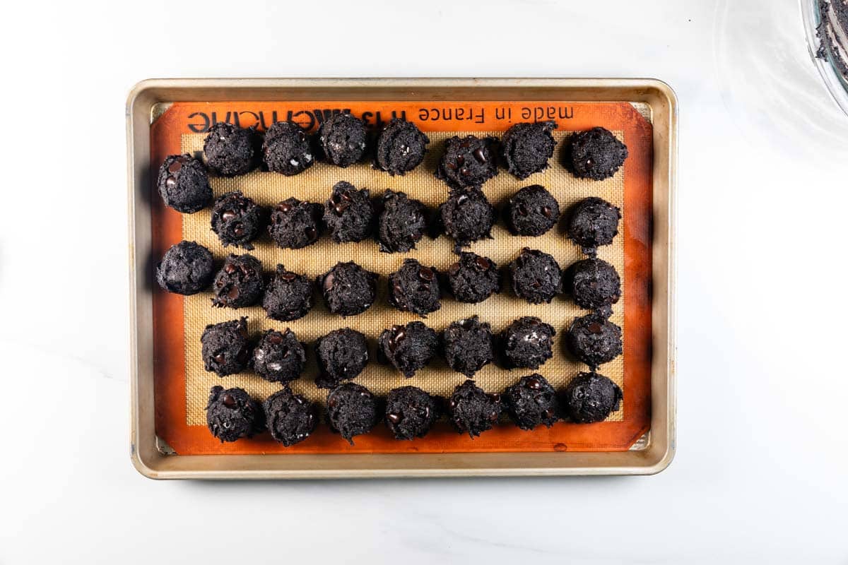 A baking sheet lined with a silicone mat holds five rows of round Chocolate Oreo Cookies dough balls, evenly spaced and ready for baking.