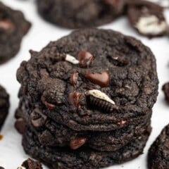 A stack of Chocolate Oreo Cookies with chocolate chips and Oreo cookie pieces, surrounded by more cookies and Oreo crumbs on a white surface.