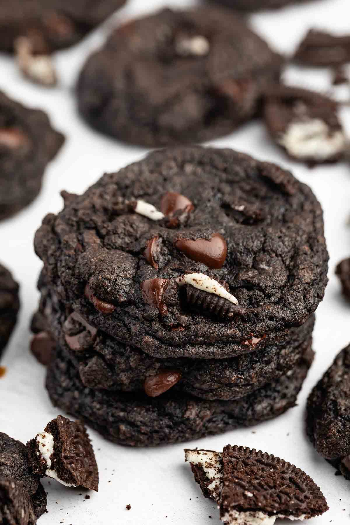 A stack of Chocolate Oreo Cookies with chocolate chips and Oreo cookie pieces, surrounded by more cookies and Oreo crumbs on a white surface.