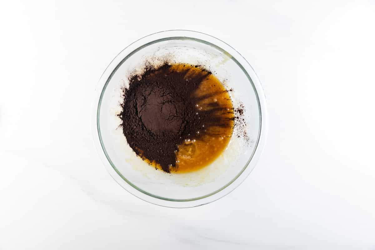 A glass mixing bowl on a white surface containing dark cocoa powder and a brown liquid mixture, partially combined for chocolate Oreo cookies.