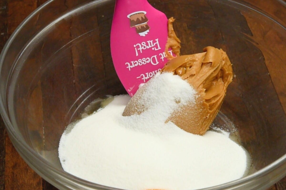 clear bowl on wood table with peanut butter sugar and egg in it and spatula.