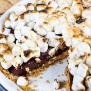 S'mores pie in white pie dish with one slice missing and ingredients around it