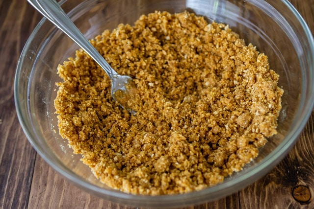 Graham cracker crust with butter in a clear mixing bowl with fork