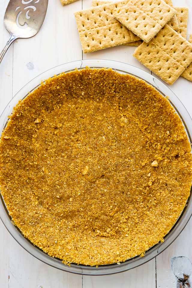 Overhead shot of graham cracker crust in clear pie dish on white wood table with graham crackers and serving silverwear
