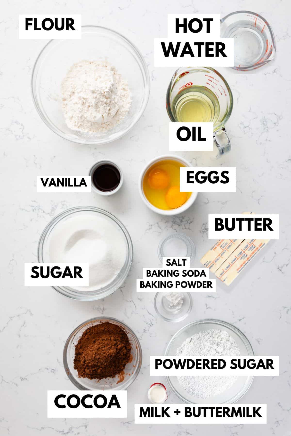 Top-down view of labeled baking ingredients in bowls and measuring cups for a chocolate cake recipe, including flour, sugar, cocoa, powdered sugar, eggs, butter, oil, hot water, milk, buttermilk, vanilla, and baking powders.