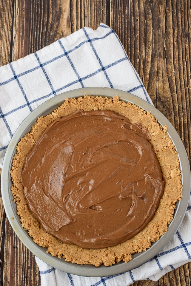 Overhead shot of pie crust and nutella later in pie dish on a wood table with white and blue checkered napkin