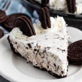 A slice of No Bake Oreo Cheesecake with a chocolate cookie crust, topped with whipped cream and whole chocolate sandwich cookies, served on a white plate. The whole pie is visible in the background.