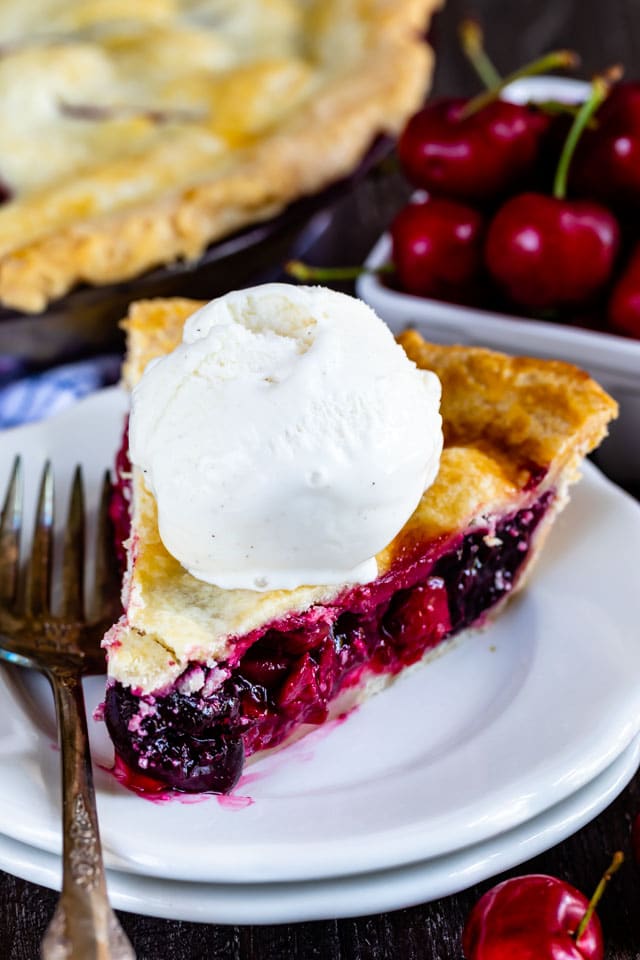 Slice of cherry pie with a scoop of ice cream on top on a white plate with fork