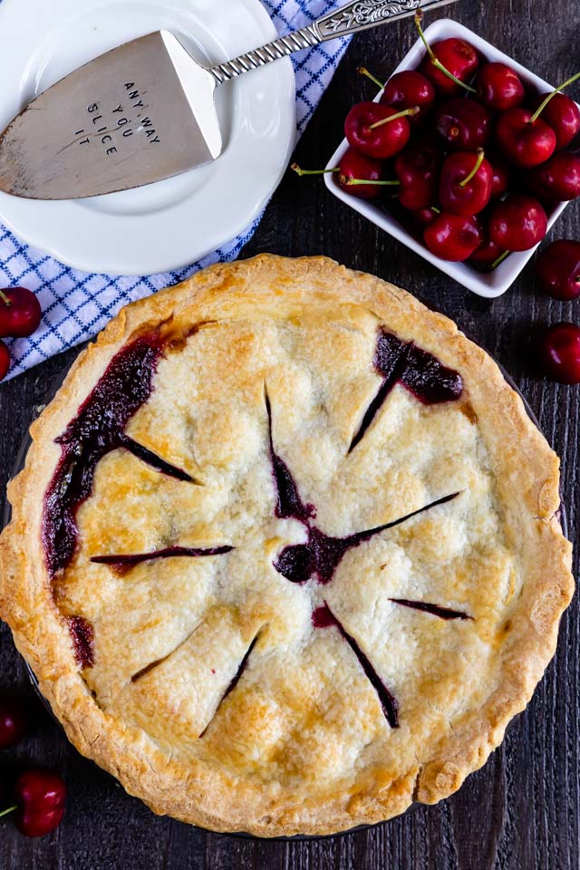 Overhead shot of full cherry pie with plate and basket of cherries on wood table