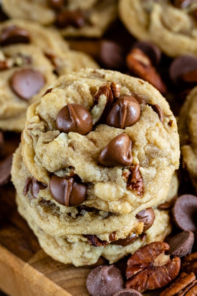 Overhead shot of chocolate chip pecan cookies stacked on a wood cutting board with chocolate chips and pecans