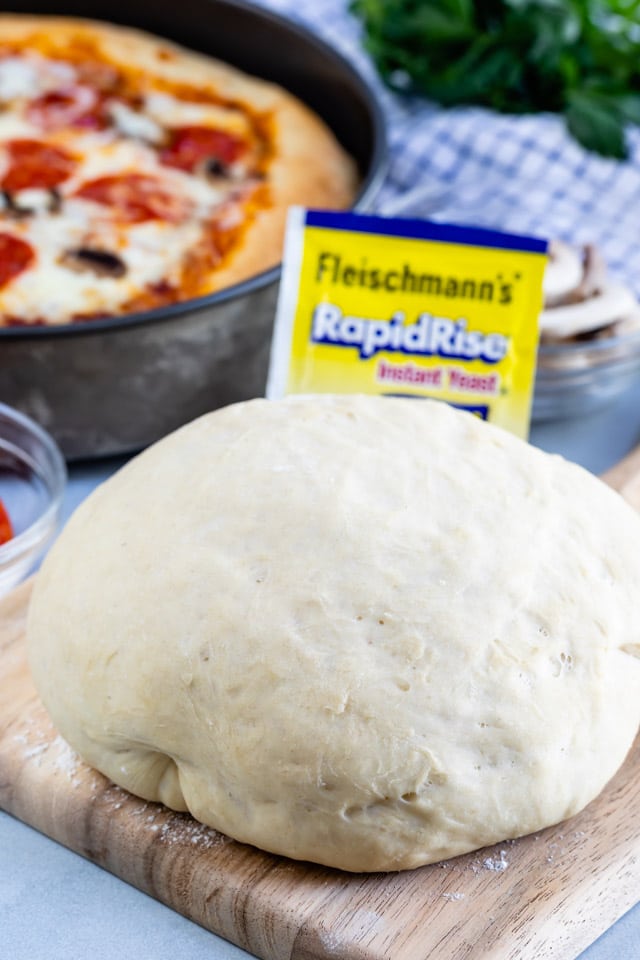 Pizza dough in a ball sitting on a wood cutting board with yeast packet and deep dish pizza in background