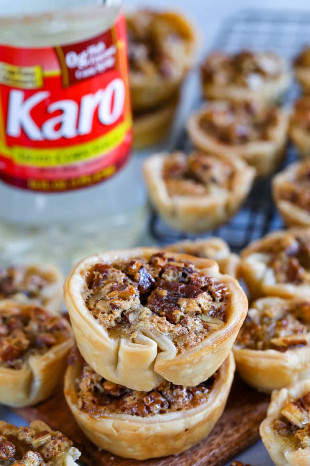 Mini pecan pies on a wood cutting board with pecans scattered around and karo corn syrup bottle in background