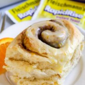 Close up of an orange sweet roll on a white plate with yeast packets behind it