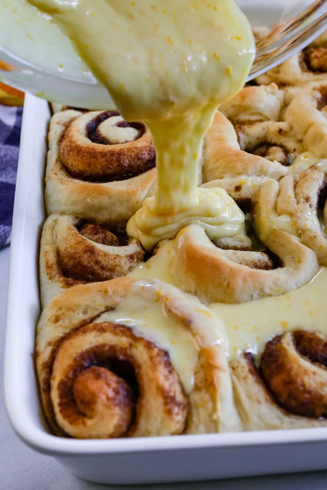 Orange sweet rolls in a white baking dish with orange icing being poured over them