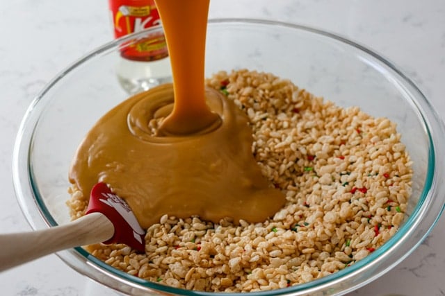 Peanut butter mixture being poured into glass mixing bowl with rice cereal and sprinkles