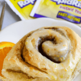 Close up photo of one orange sweet roll on a white plate with yeast packets in background with recipe title on top