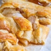 Close-up of a golden, flaky Apple Strudel braid with a shiny glaze on top and a sweet filling oozing between the folds, resting on a piece of parchment paper.