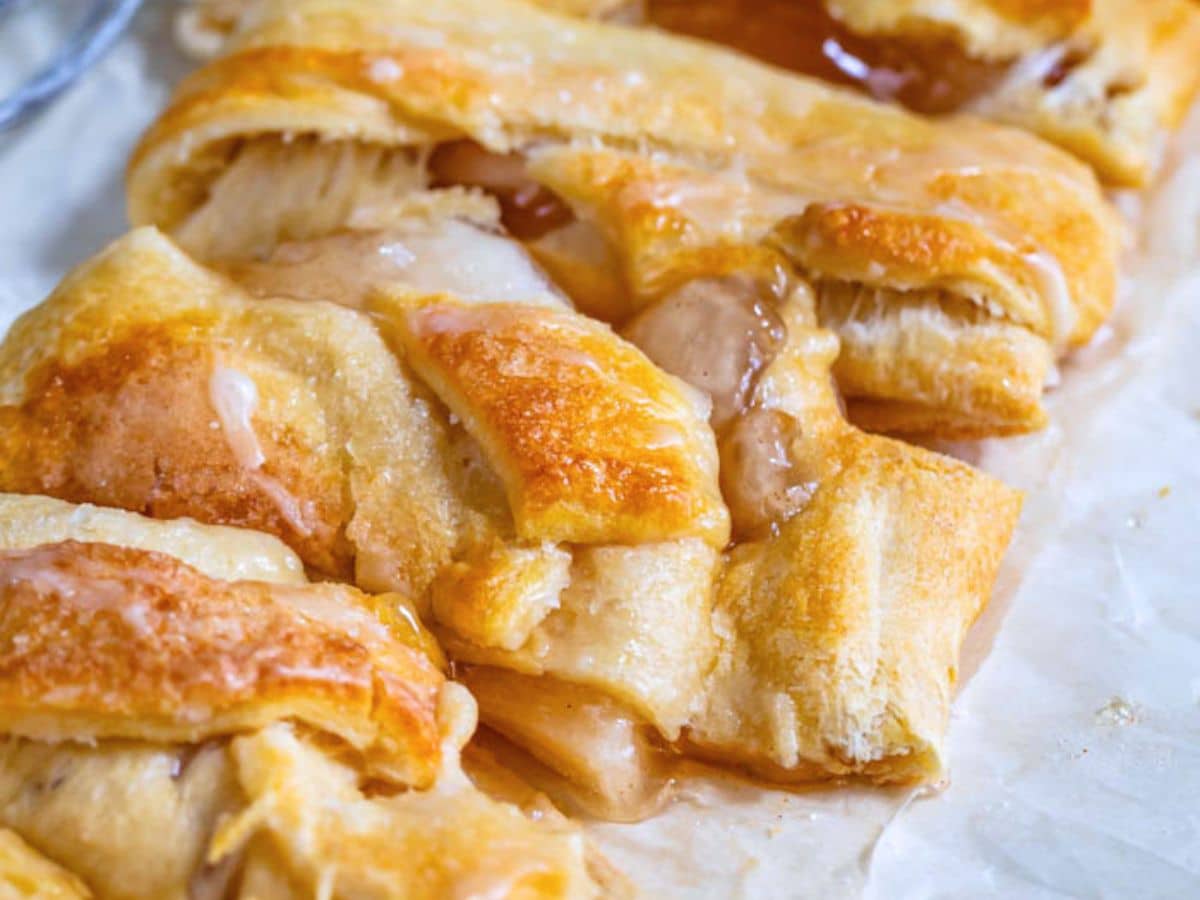 Close-up of a golden, flaky Apple Strudel braid with a shiny glaze on top and a sweet filling oozing between the folds, resting on a piece of parchment paper.