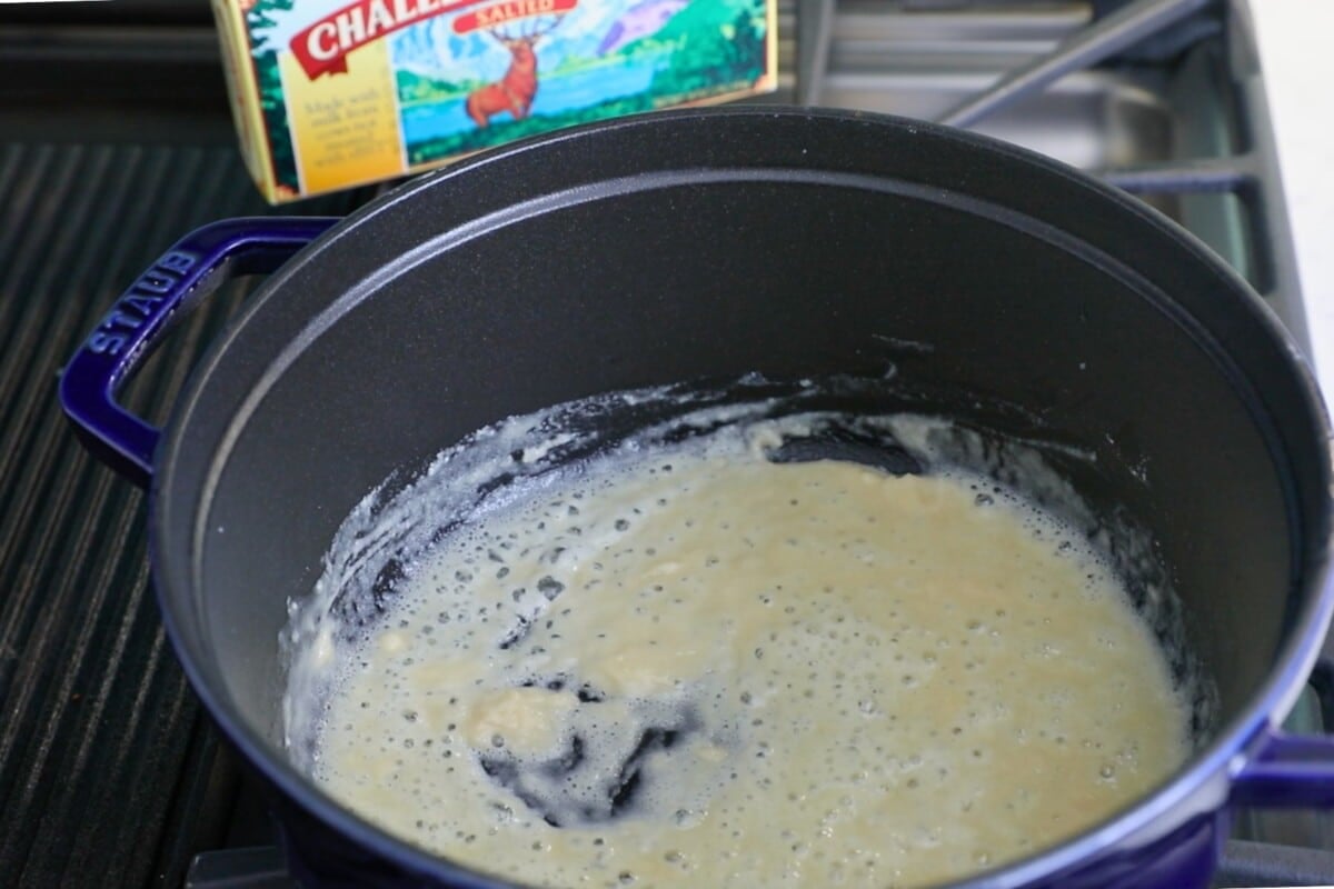 A blue pot on a stove contains a bubbling mixture of butter and flour, hinting at the beginnings of a cheesy chicken casserole. In the background, a box of Challenge butter stands proudly as the roux thickens, ready to create rich sauces for your savory creation.