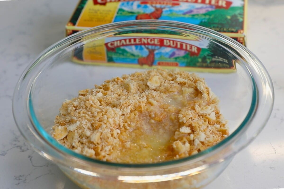 A glass bowl brims with a mixture of melted butter and crushed crackers, blending into a crumbly texture for the perfect cheesy chicken casserole. Behind the bowl, a partially visible box of Challenge Butter rests on a marble surface, hinting at delicious possibilities.