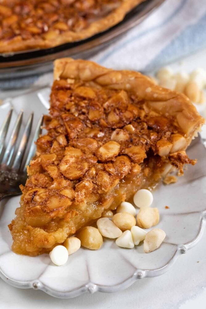 One slice of macadamia nut pie on a white scalloped plate with extra white chocolate chips and macadamia nuts