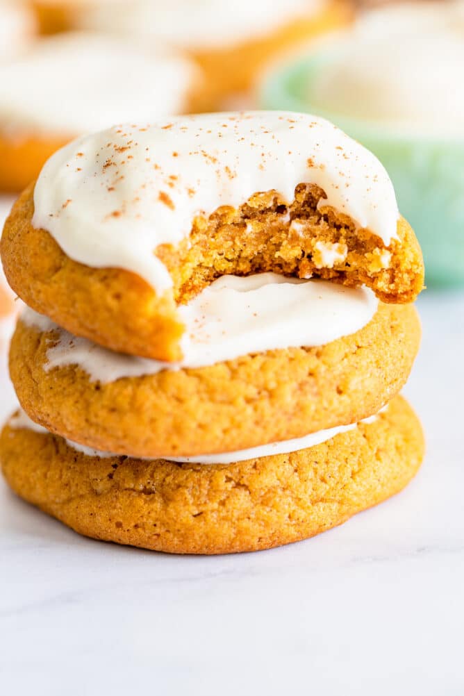 Stack of three pumpkin cookies topped with cream cheese frosting and pumpkin spices with a bite missing out of one cookie