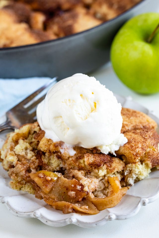 Snickerdoodle apple cobbler on a white plate with vanilla ice cream scoop on top and green apple in background