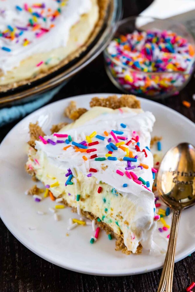 Slice of funfetti pie on a white plate with silver spoon and sprinkles in the background