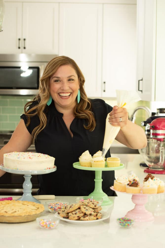 woman holding piping bag frosting cupcakes with desserts in front of her