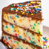 One slice of layered funfetti birthday cake with chocolate frosting and rainbow sprinkles on a white plate with fork and recipe title on image