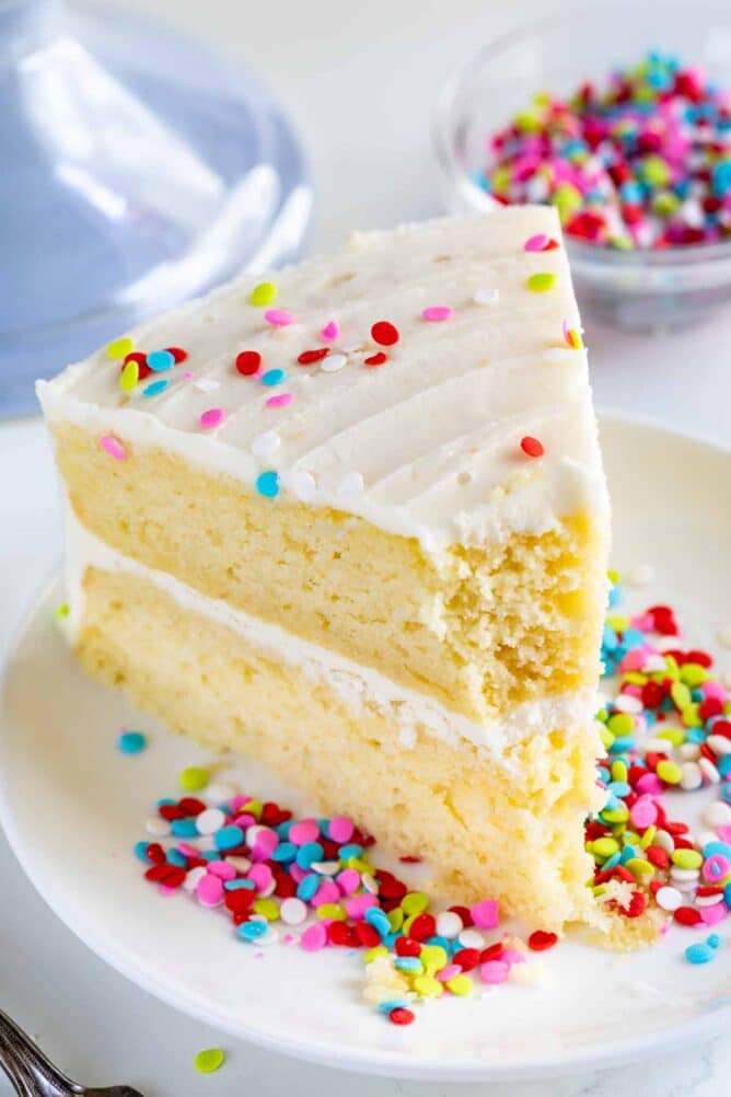 In primo piano di una fetta di torta gialla a strati con glassa alla vaniglia e sprinkles arcobaleno con un morso missing