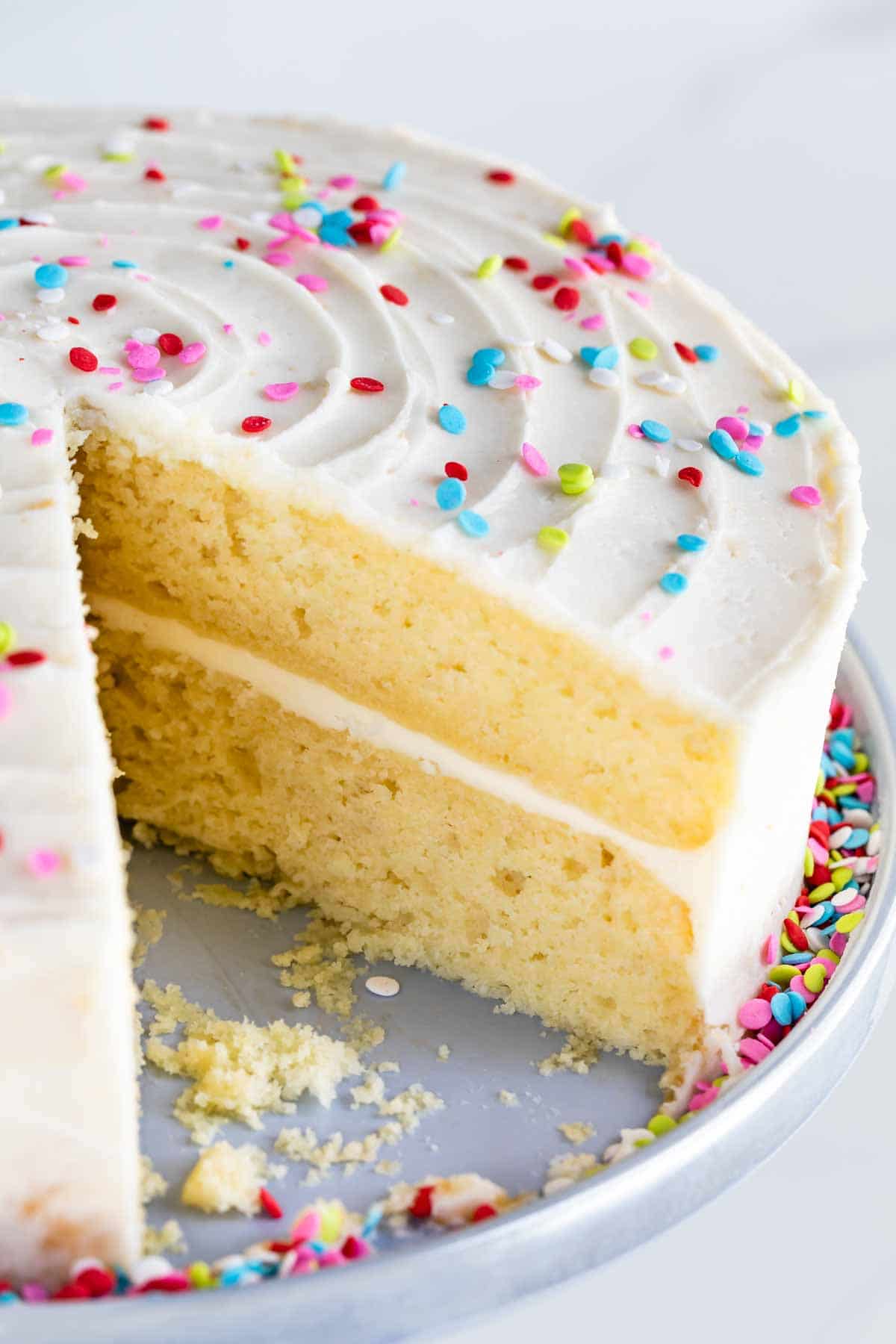 Yellow layered cake on a cake stand with vanilla buttercream and rainbow sprinkles and one slice missing