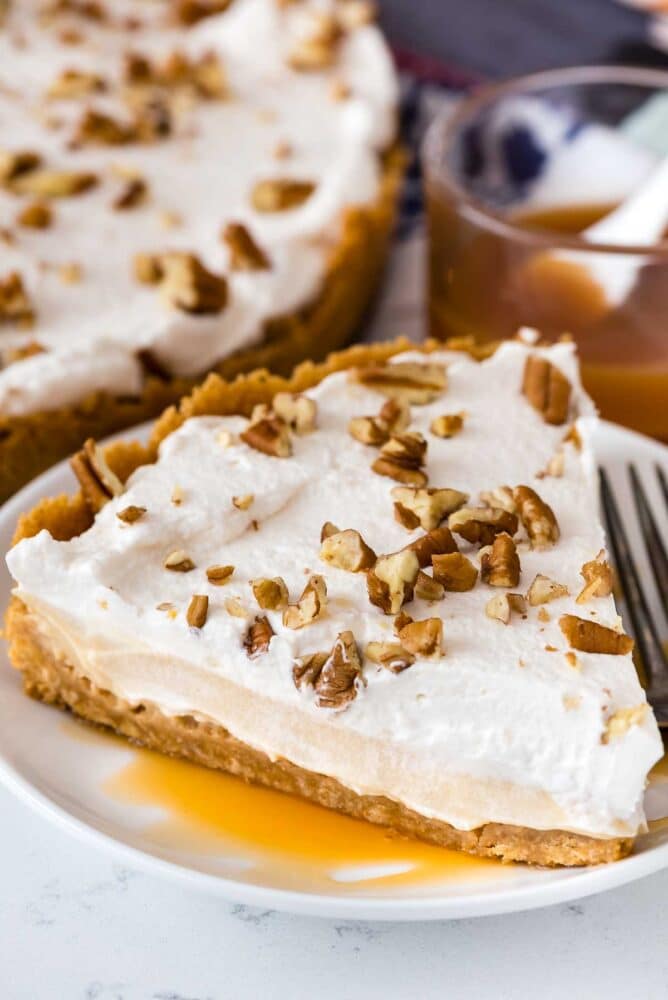 A slice of butterscotch pie with pecans on top on a white plate with rest of pie in background