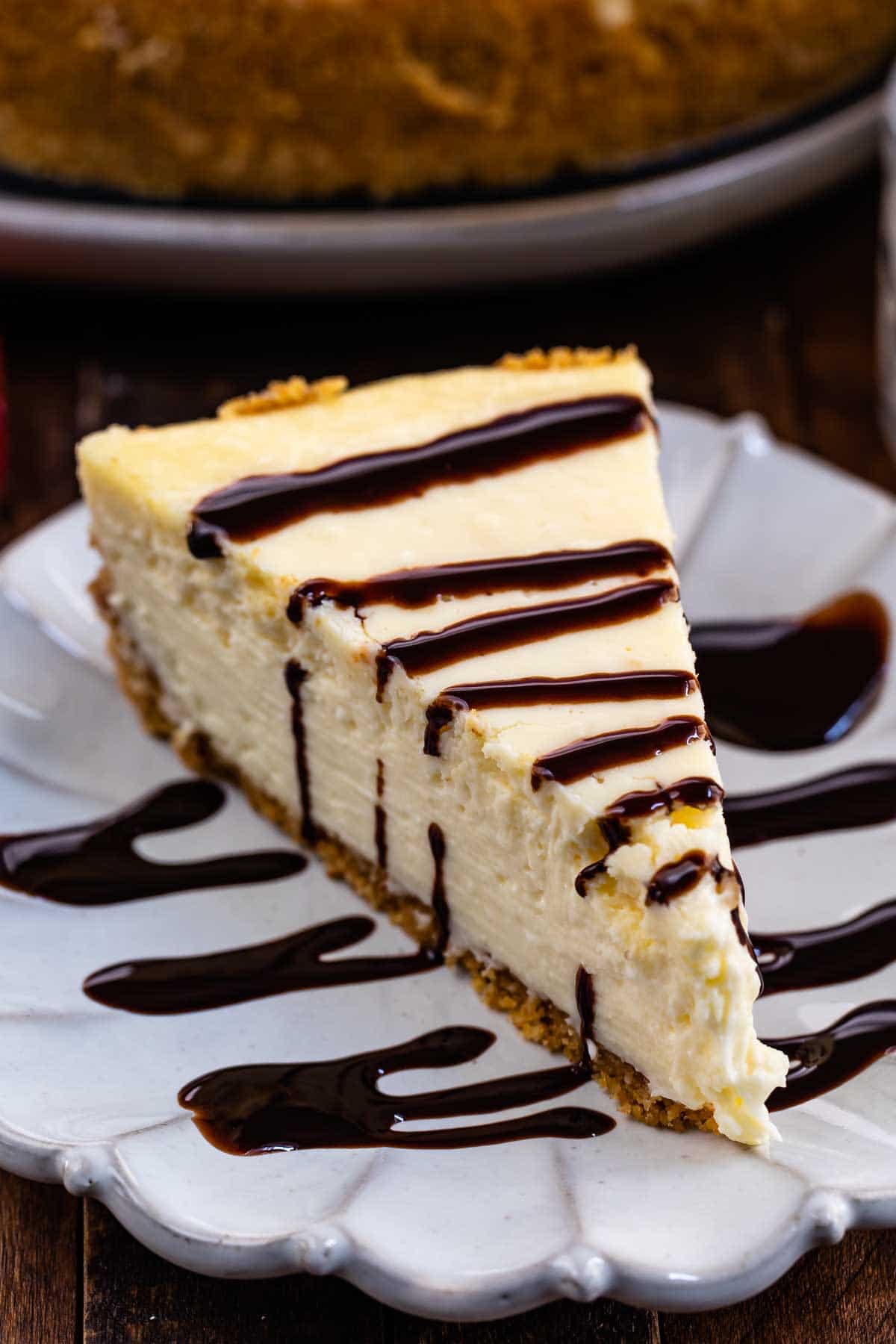One slice of classic cheesecake on a scalloped plate with a chocolate sauce drizzle