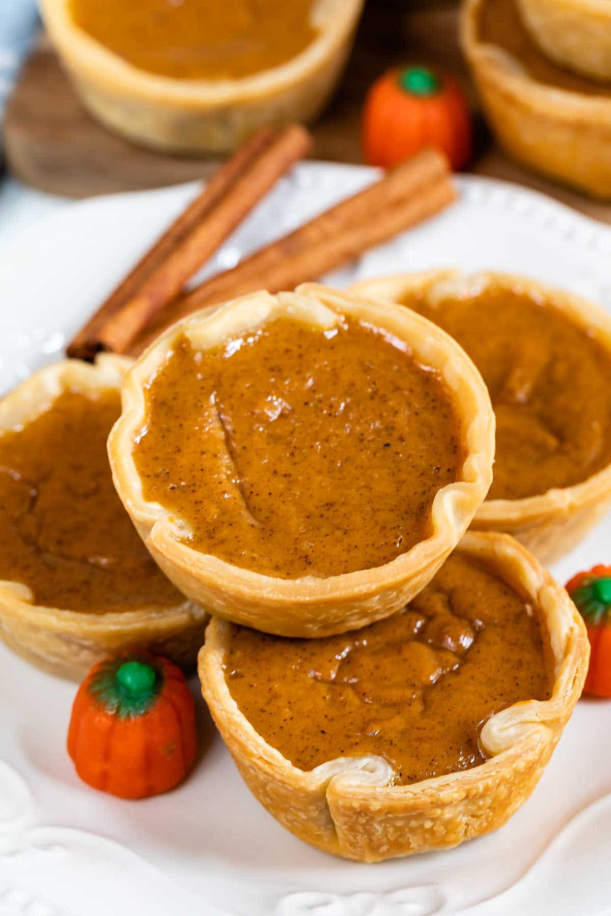 Mini pumpkin pies stacked on top of eachother on a white plate with pumpkin candy and cinnamon sticks for decoration