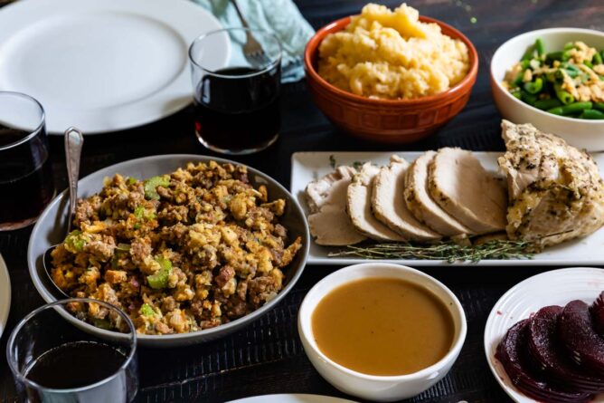 table with place settings and sausage stuffing, sliced turkey breast, bowl of gravy, cranberry sauce, orange bowl of mashed potatoes and bowl of green beans with glasses of coke