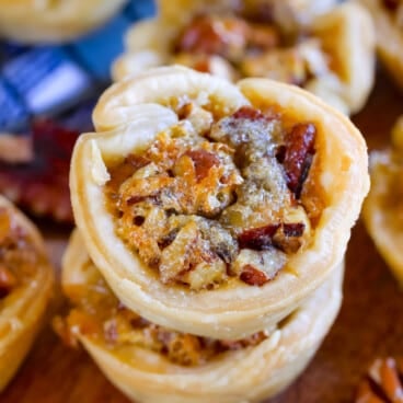 Close-up of stacked mini pecan pies on a wooden surface, showing flaky pastry filled with caramelized pecans. Out-of-focus tarts and a cooling rack appear in the background.