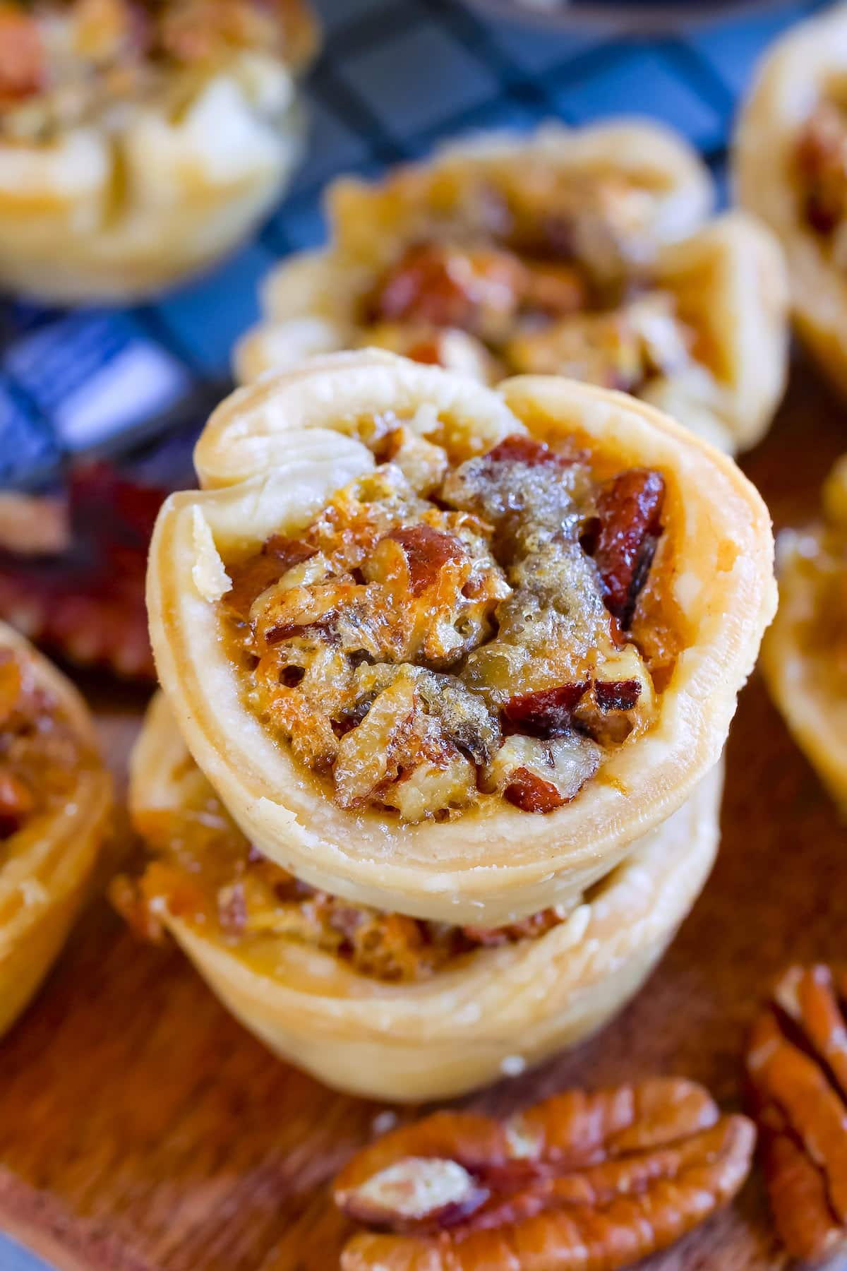 Close-up of stacked mini pecan pies on a wooden surface, showing flaky pastry filled with caramelized pecans. Out-of-focus tarts and a cooling rack appear in the background.