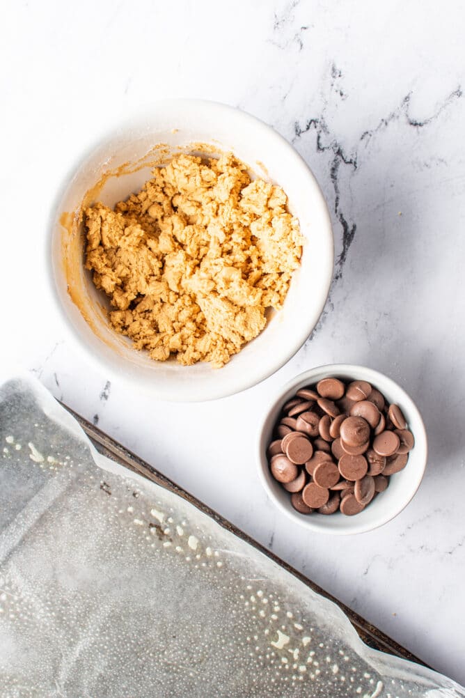 Overhead view of peanut butter ball mixture and chocolate in separate dishes next to sheet pan