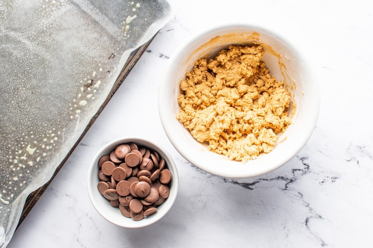 Overhead view of peanut butter ball mixture and chocolate in separate dishes next to sheet pan