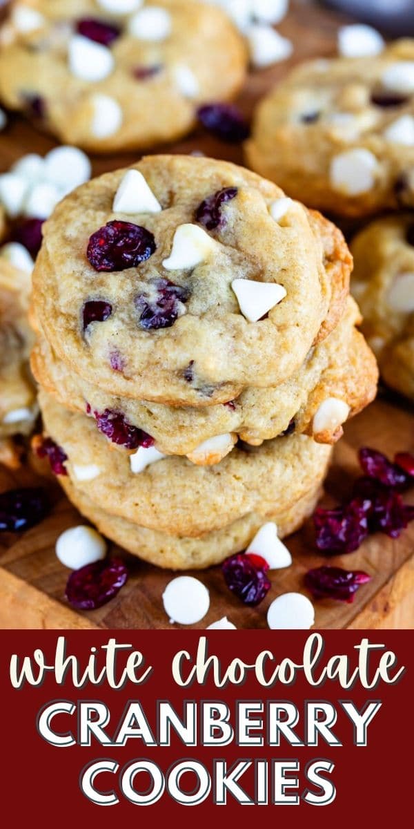 Stack of cranberry white chocolate cookies on a wood cutting board surrounded by cranberries and white chocolate chips and recipe title on bottom of image