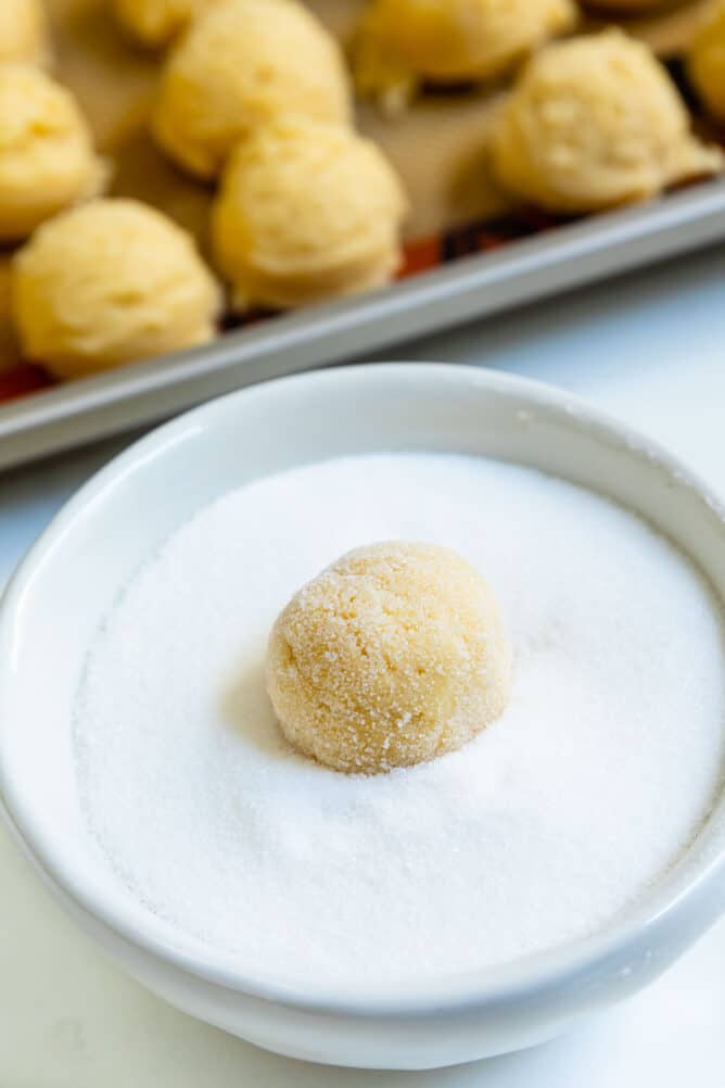 One peppermint blossom cookie dough ball being rolled in sugar with more cookie dough balls in background