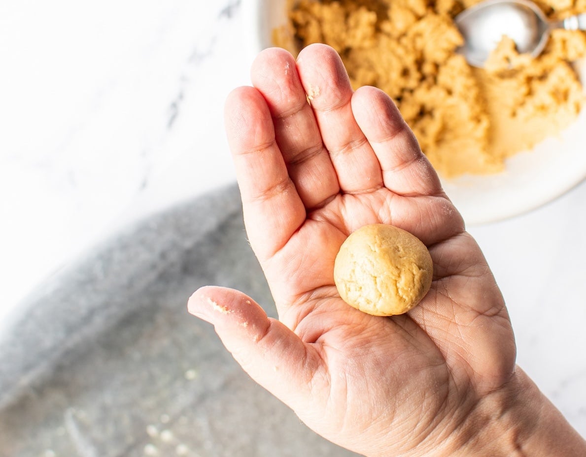 A person's palm holds a small, round ball of beige cookie dough—perfect for making a classic peanut butter balls recipe. In the background, a white bowl filled with more dough and a metal spoon sits on a marble surface.