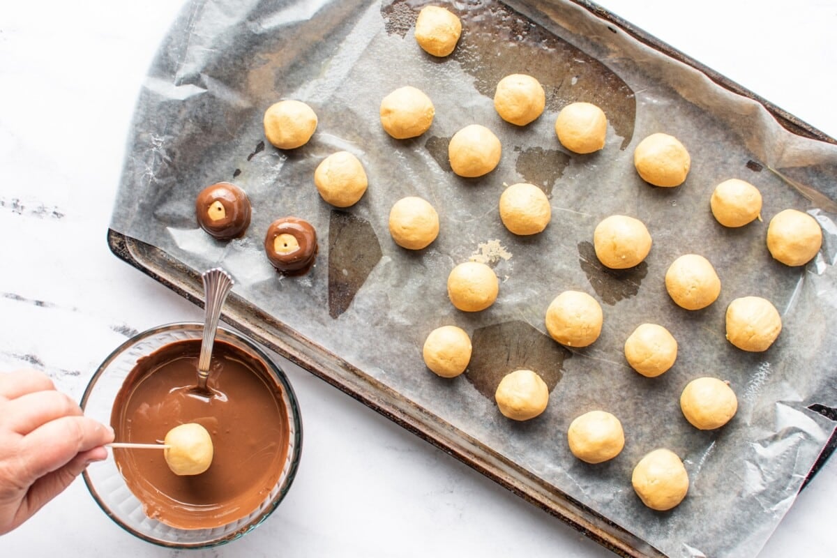 A hand dips a peanut butter ball into melted chocolate, showcasing a key step in this irresistible peanut butter balls recipe. Nearby, a baking tray holds rows of undipped and partially dipped peanut butter balls on parchment paper.